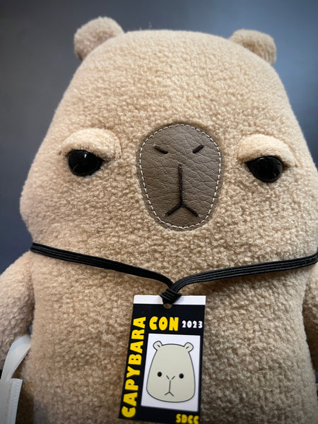 Unimpressed and Judgy looking Capybara plush with event badge and a tote bag accessories - face front close-up